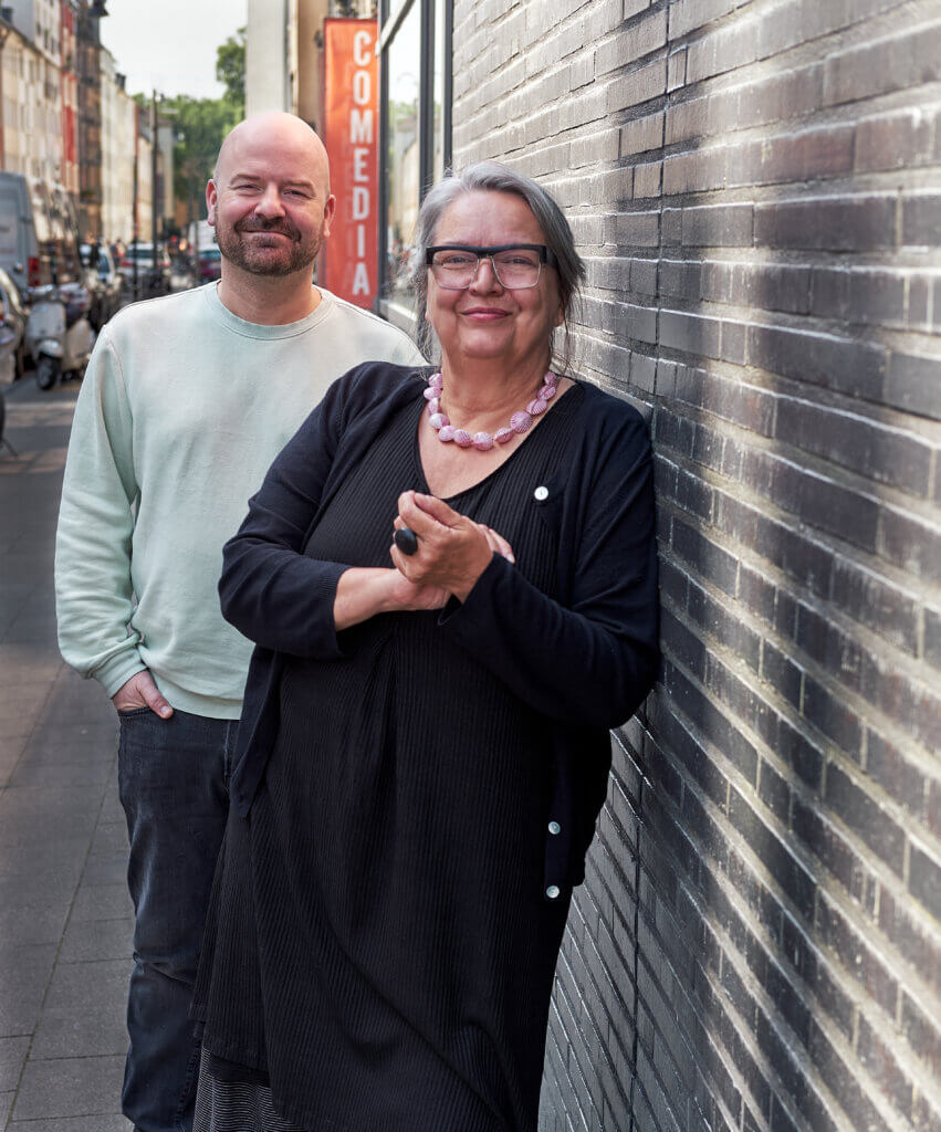 Manuel Moser, Leiter der Abteilungfür junges Publikum und Jutta M. Staerk, Leiterin, stehen an einer Hauswand gelehnt hintereinander.