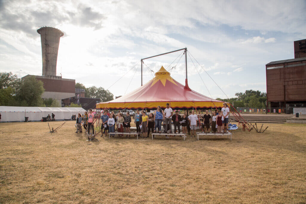 Impression vom europefiction-Camp 2019 auf der Wiese am Theater. Ein Zirkuszelt mit rot-gelbem Dach, davor stehen sehr viele Menschen in einer Reihe.