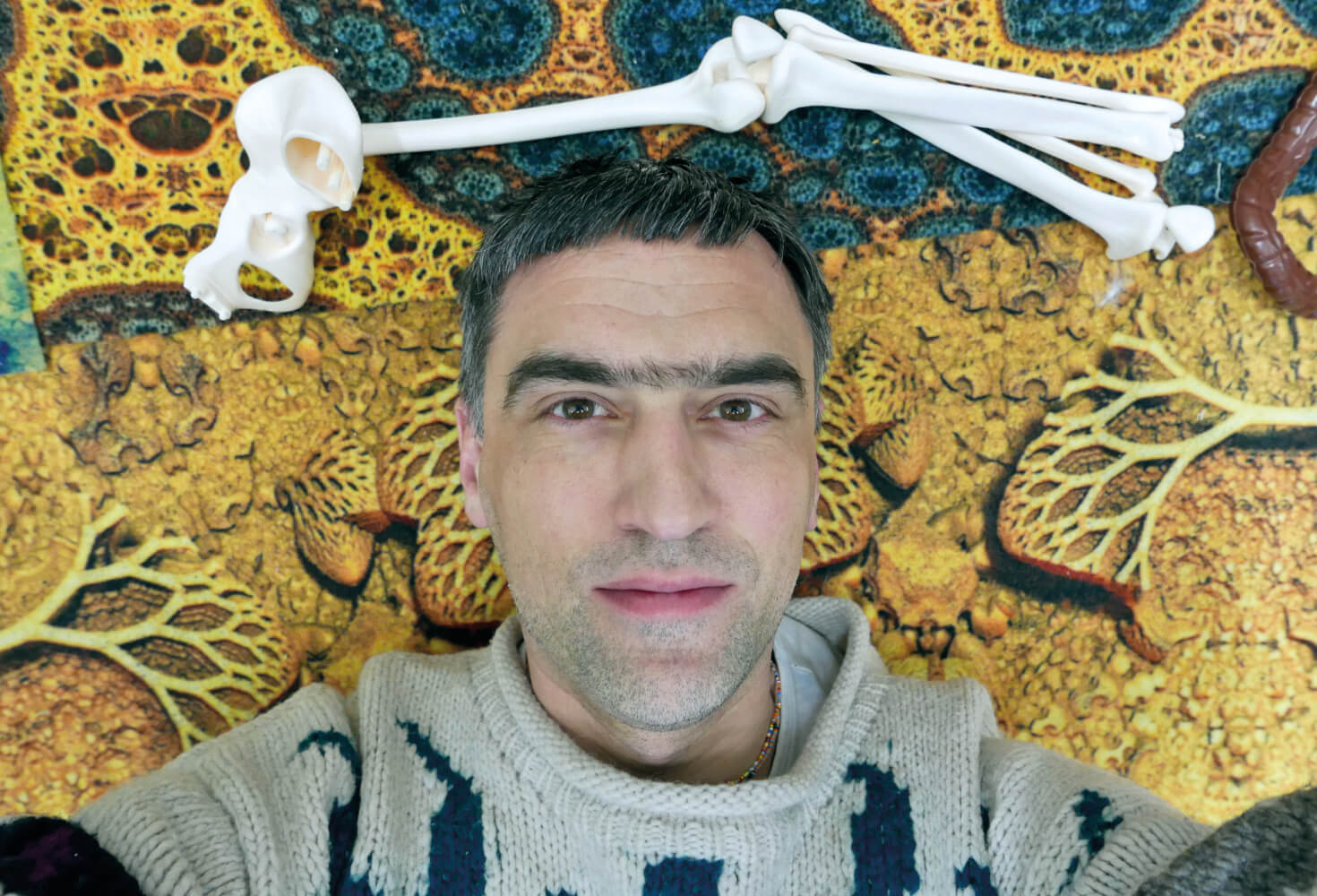 Selfie von Markus Selg vor einer strukturierten, gold-blauen Wand, über ihm einzelne weiße Knochen.