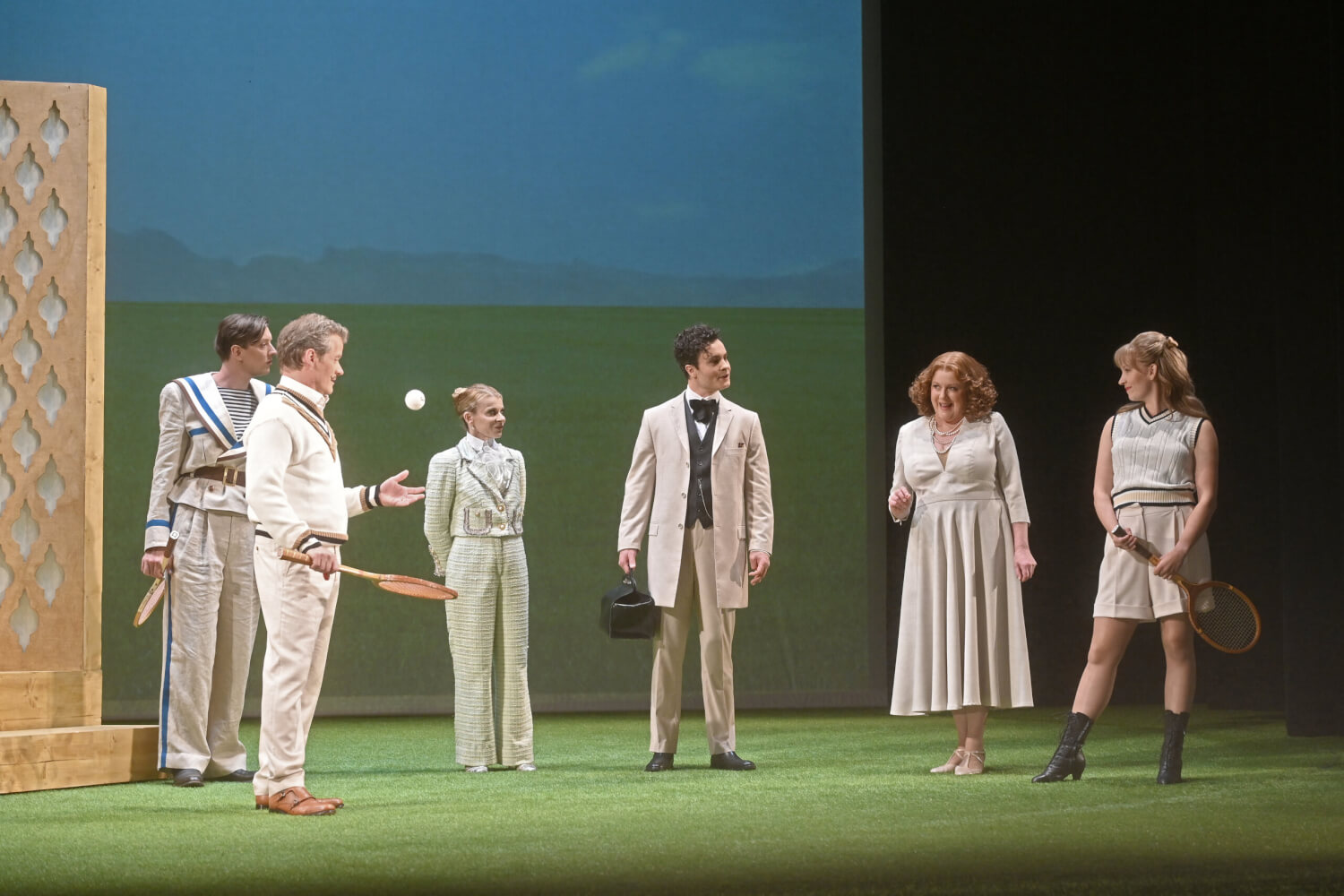Aufführungsfoto von „Das weite Land“ von Arthur Schnitzler am Theater Baden-Baden. Drei Männer und drei Frauen stehen in beige-farbener Kleidung auf einem kurzen Kunstrasen. Ein Mann hat einen alten Holztennissschläger in der Hand und wirft einen Tennisball hoch. Ein anderer Mann und eine Frau haben ebenfalls Tennisschläger in der Hand und ein Mann einen schwarzen Arztkoffer.