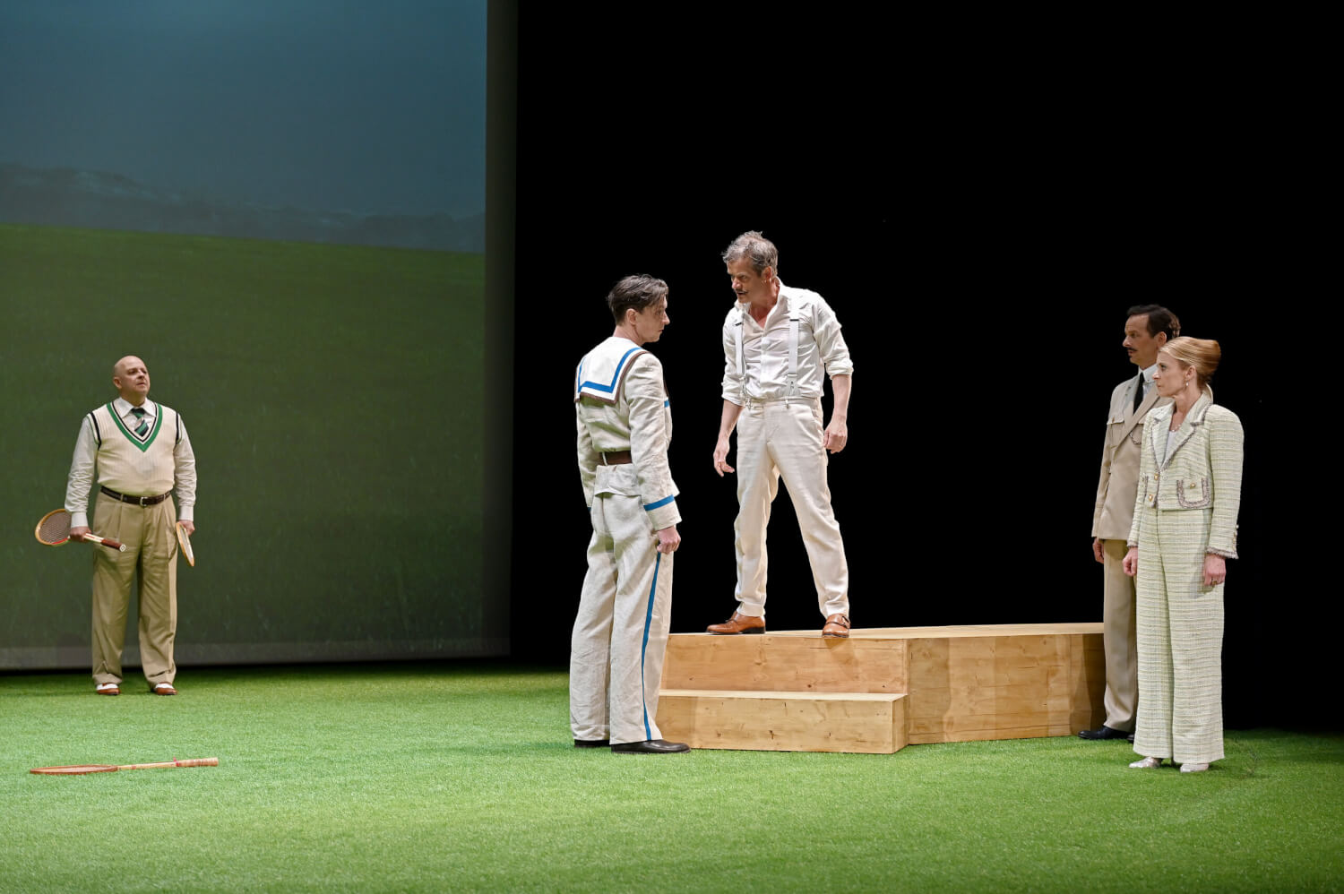 Aufführungsfoto von „Das weite Land“ von Arthur Schnitzler am Theater Baden-Baden. Auf einem kurzen grünen Kunstrasen stehen vier Männer und eine Frau in beiger Kleidung. Ein Mann steht auf einem Holzpodest mit zwei Stufen und blickt einen jungen Mann streng an, der zur Seite schaut.