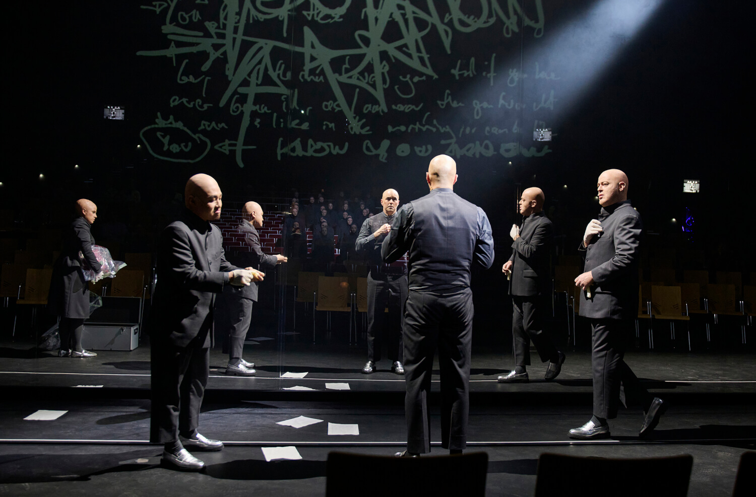 Aufführungsfoto von „WE (WIR)“ von Sarah Nemtsov am Theater Dortmund. Sieben Männer in schwarzen Anzügen und Glatzen stehen auf einer stufigen Bühne.