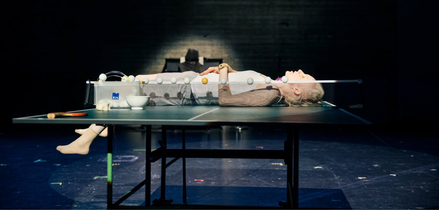 Aufführungsfoto von „Man kann auch in die Höhe fallen“ nach Joachim Meyerhoff am Badischen Staatstheater Karlsruhe. Eine Frau liegt auf einer Tischtennisplatte, ihre Beine lässt sie herunterhängen, die Arme ruhen auf dem Bauch, vor ihr auf dem Netz liegen mehrere Tischtennisbälle.