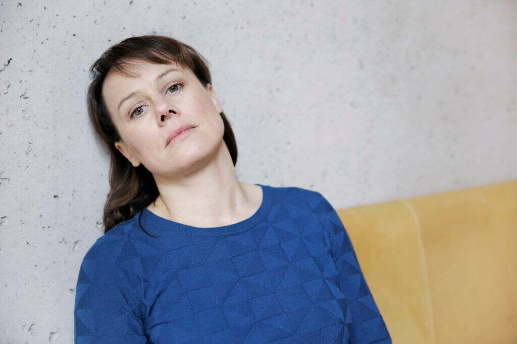 Porträt von Eva Löbau. Sie hat den Kopf an eine Betonwand angelehnt und trägt einen blauen Pullover.