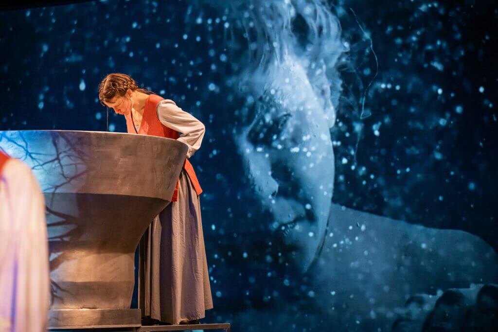 Aline Bucher beugt sich über einen Brunnen. Im Hintergrund wird ihr Gesicht auf die Bühnenwand projiziert. Ihre Verzweiflung ist spürbar.