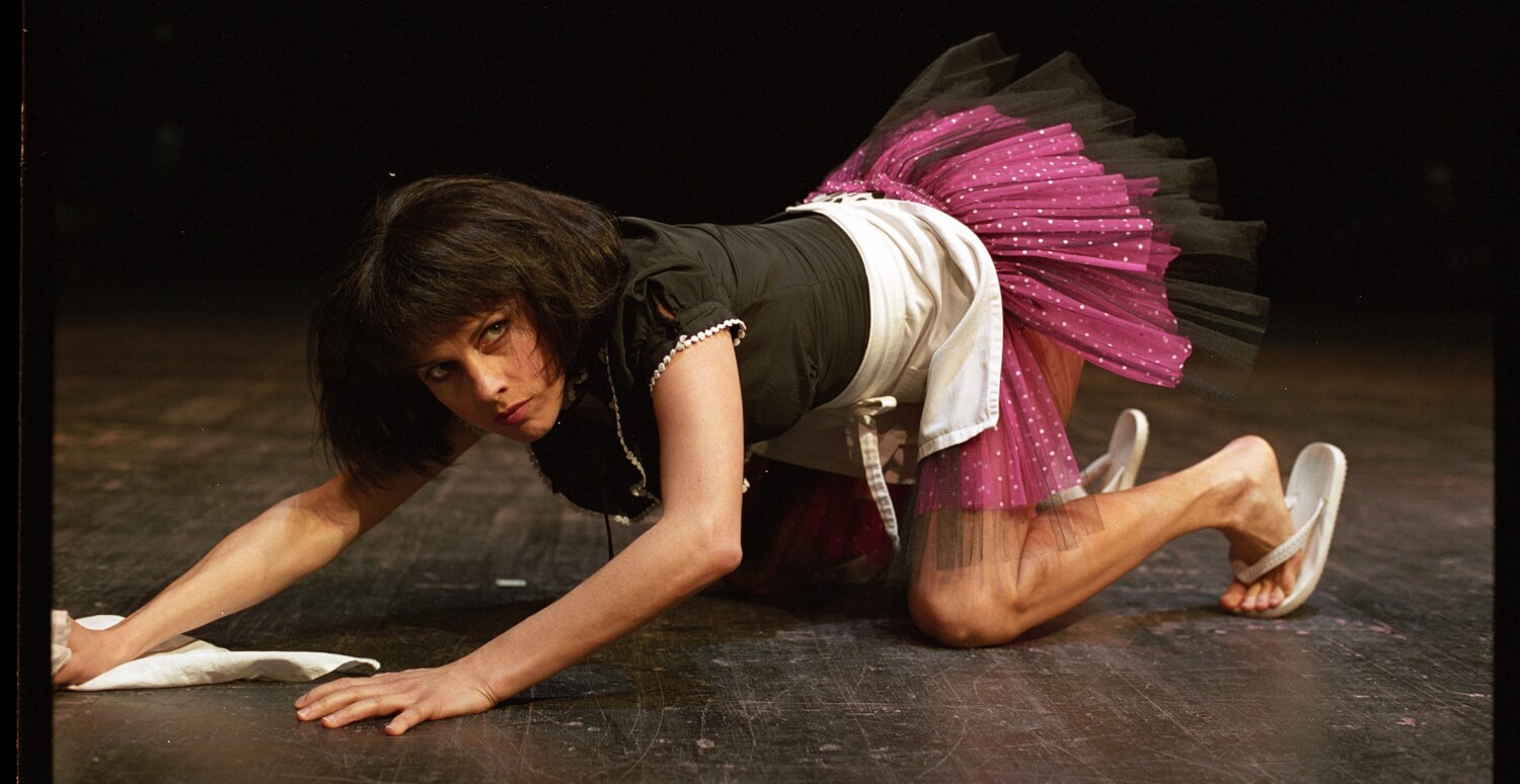 Aufführungsfoto von Dimiter Gotscheffs „Tartuffe“-Inszenierung. Die Schauspielerin Judith Rosmair kniet mit Tütü, einem schwarzen, engen T-Shirt und weißen Flip Flops auf dem schwarzen Bühnenboden mit einem weißen Putztuch in der rechten Hand.