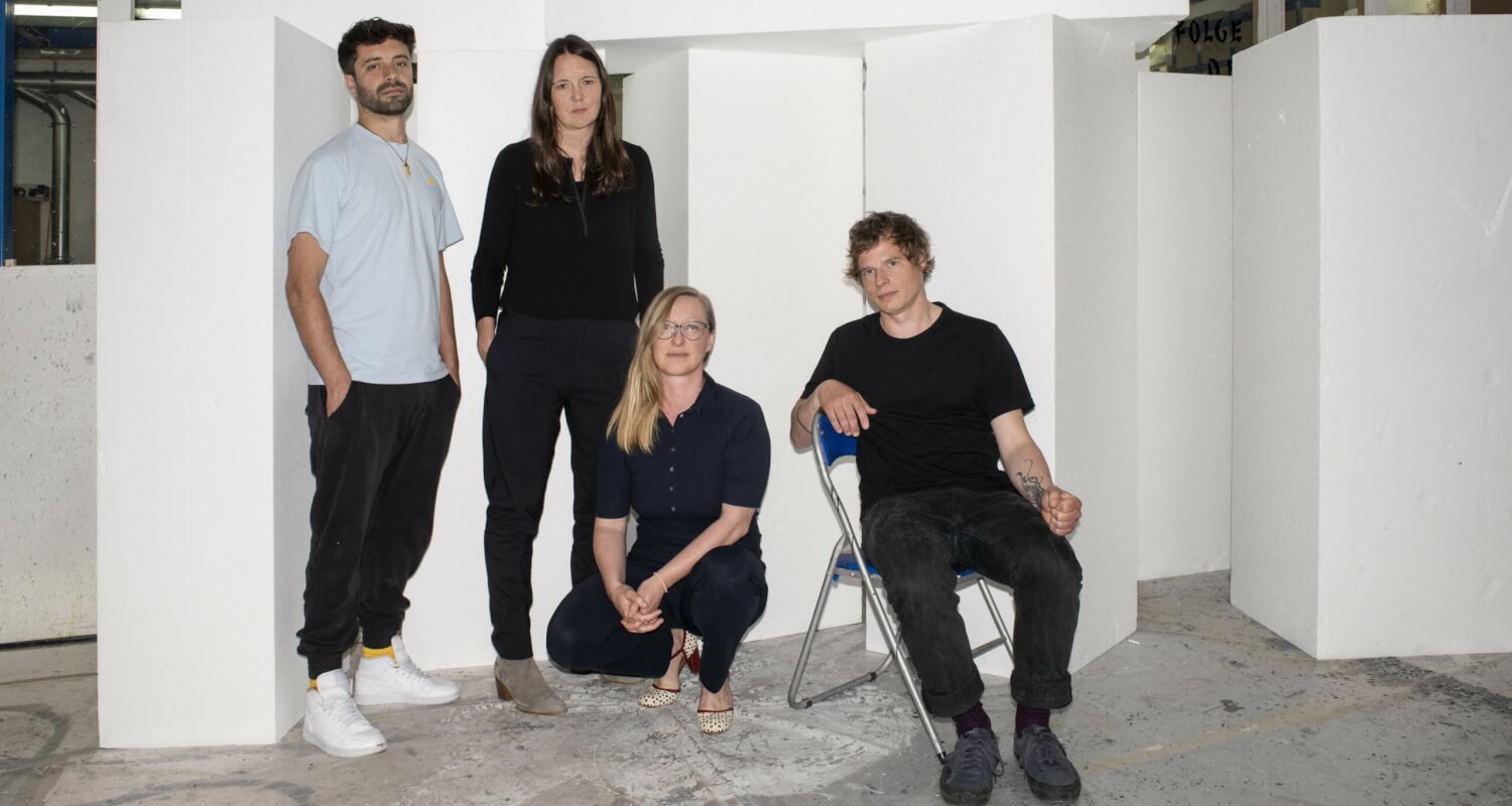 Antú Romero Nunes, Inga Schonlau, Anja Dirks und Jörg Pohl (v. l. n. r.). In einem Raum mit niederiger Decke und weißen Wänden stehen ein Mann und eine Frau links, in der Mitte hockt eine Frau und recht sitzt ein Mann auf einem Klappstuhl.