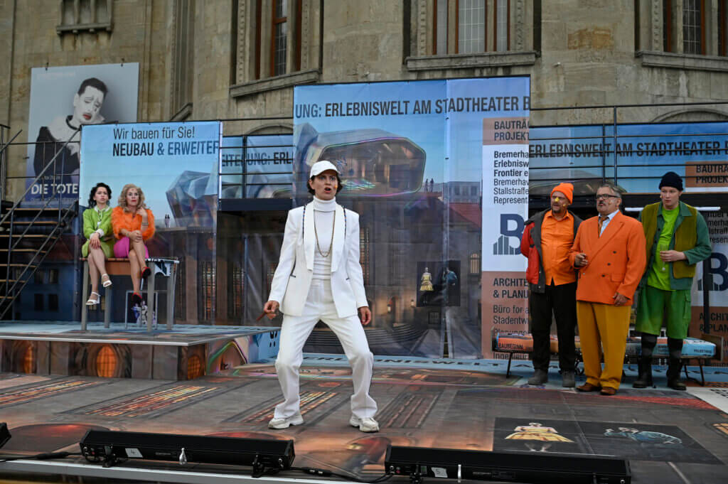 Der Revisor steht in weißer Kleidung, Kappe und Goldkettchen breitbeinig auf der Bühne. Im Hintergrund steht die Stadtgesellschaft und starrt ihn an.