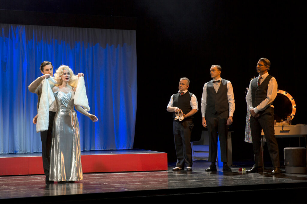Marylin steht links im glitzernen Abendkleid. Ihr wird eine Felljacke übergestreift. Rechts stehen die Tritones.