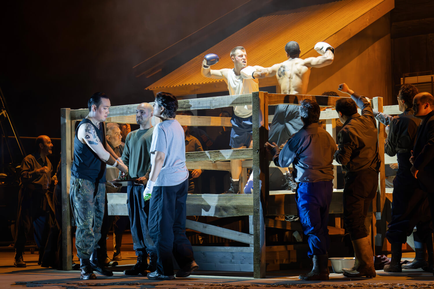 Aufführungsfoto von „Tancredi“ von Gioachino Rossini an der Oper Frankfurt. Zwei Boxer holen zum Schlag in einem hölzernen Boxring zum Schlag gegeneinander aus. Drumherum stehen Männer und feuern sie an.