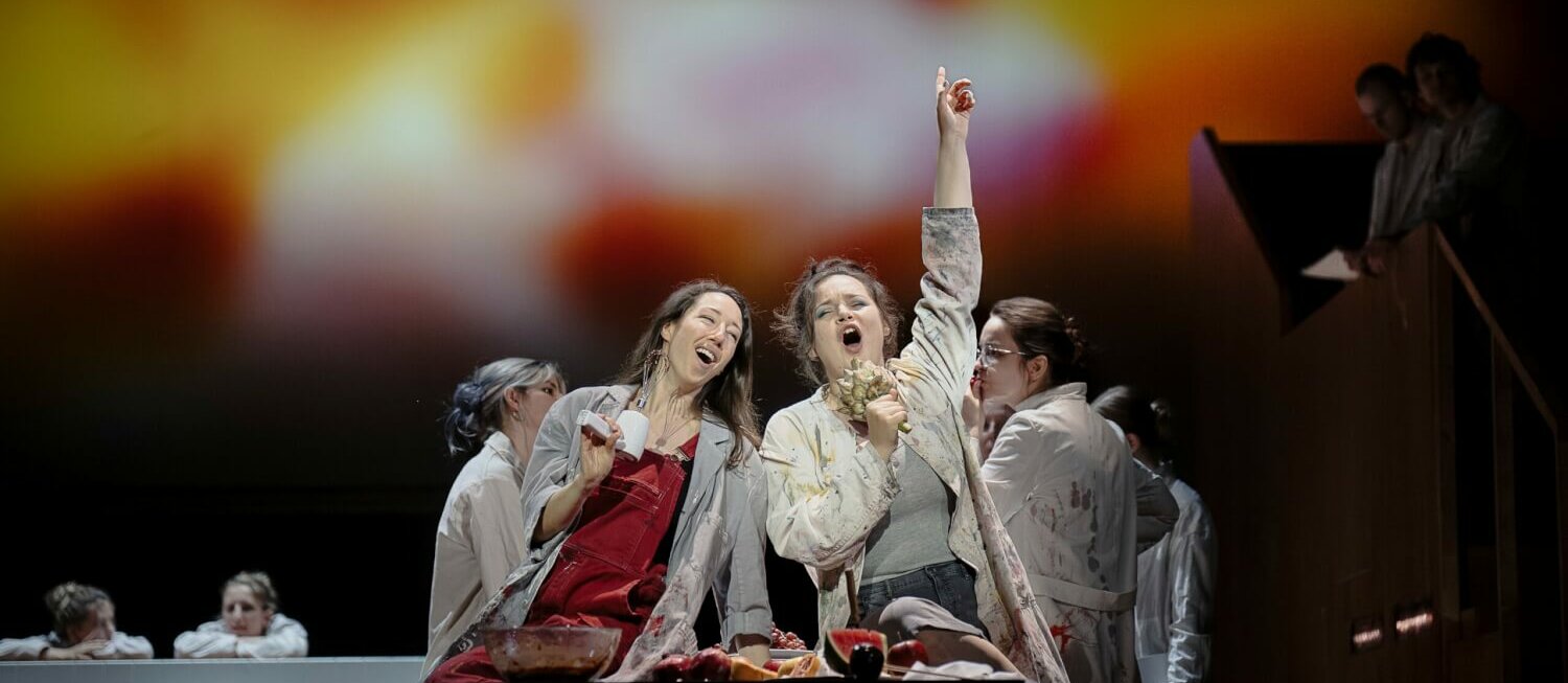 Camille Sherman und Lena Geiger sitzen nebeneinander. Beide singen fröhlich. Lena Geiger erhebt triumphal die Faust zum Himmel. In ihrer linken hält sie eine Artischocke.