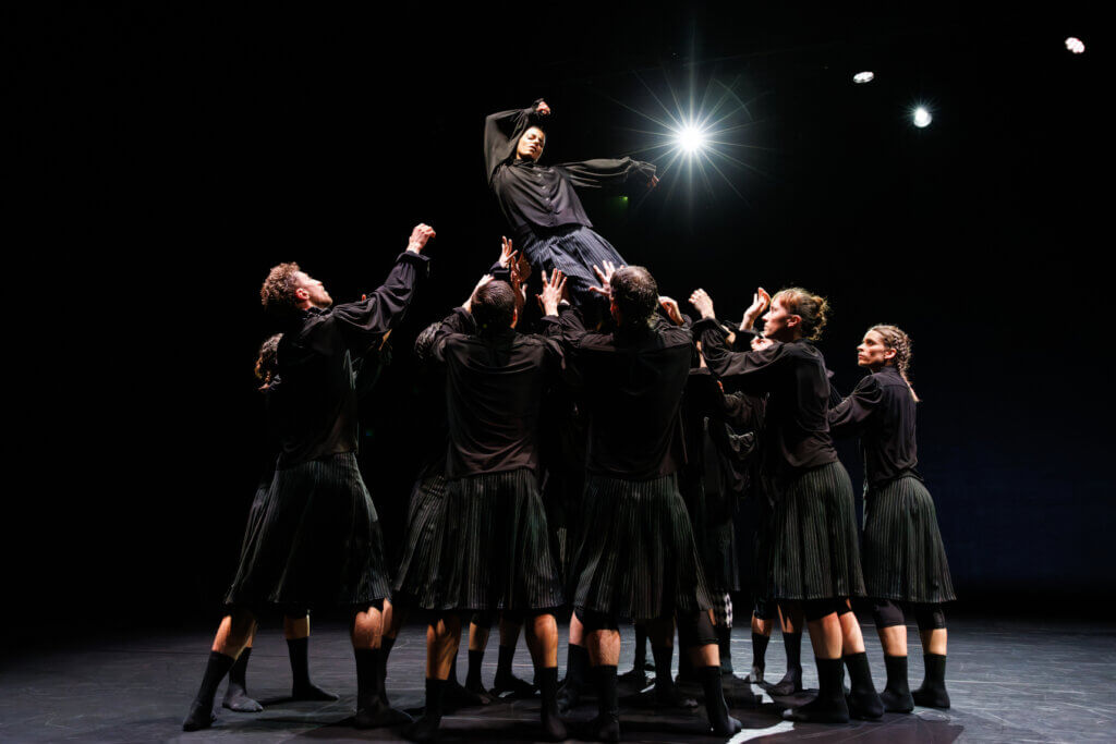 Aufführungsfoto von „Luck“ von Aszure Barton am Theaterhaus Stuttgart. Tänzerinnen und Tänzer in schwarzen Faltenröcken und schwarzen Blusen fangen eine Tänzerin auf, die in ihrer Mitte nach unten sinkt.