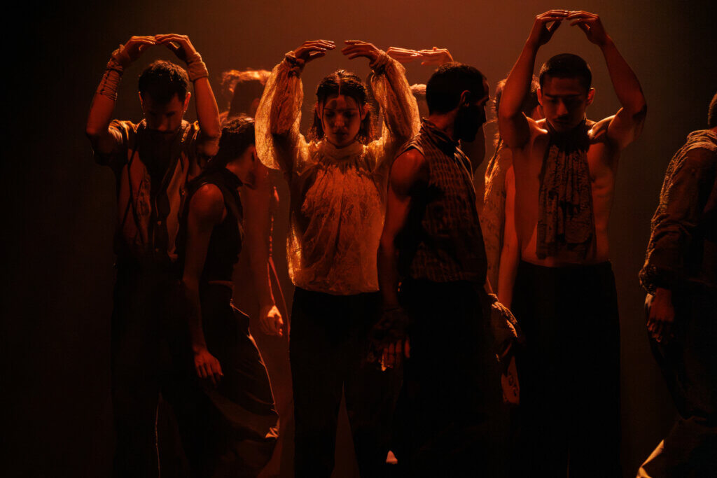 Aufführungsfoto von „Any way the wind blows“ von Hofesh Shechter am Theaterhaus Stuttgart. Tänzerinnen und Tänzer steht dicht zusammen, Blick nach unten gerichtet, die Arme über den Köpfen in orange-rotem Dämmerlicht.