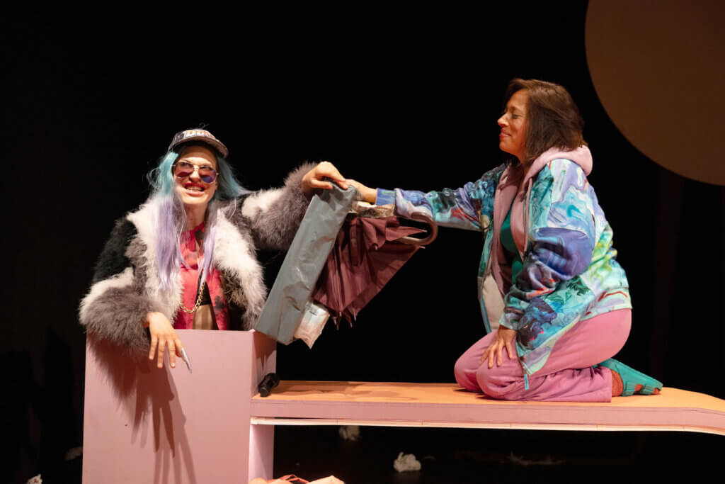 Aufführungsfoto von „Milch und Schuld“ von Sina Ahlers am Theater Lübeck. Eine Frau Mit Sonnenbrille, dickem Pelzmantel, ausgewaschenen blau-lila Haaren und Schirmmütze sitzt in einem engen Quadrat. Und hält der Frau neben ihr, die hockt, einen zusammengefalteten Regenschirm entgegen.