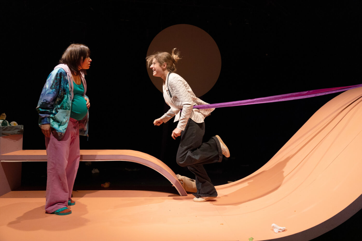 Aufführungsfoto von „Milch und Schuld“ von Sina Ahlers am Theater Lübeck. Eine schwangere Frau schaut eine andere Frau an, die am Bauch gehalten von einem Theraband ihr entgegenläuft. Die Bühne ist geschwungen.