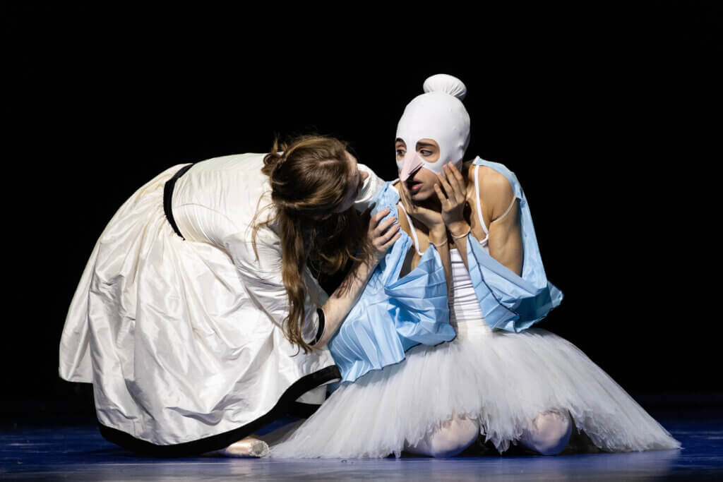 Aufführungsfoto von „Wunderland“ von Alexei Ratmansky des Hamburg Ballett an der Staatsoper Hamburg. Eine Frau mit weißem Tütü und einer Strumpfmaske über den Kopf hockt auf dem Boden, neben ihr eine andere Frau, die sich zu ihr runterbeugt und sie anschaut.