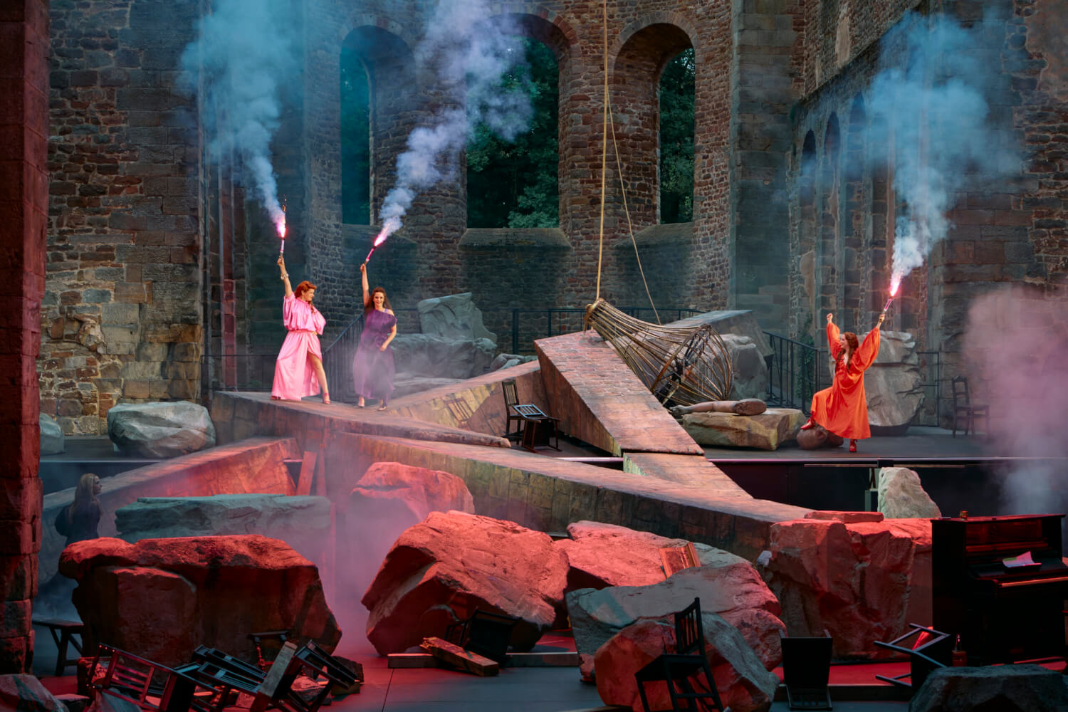 Aufführungsfoto von „Lysistrata“ von Amanda Lasker-Berlin nach Aristophanes bei den 75. Bad Hersfelder Festspielen. Massive Felsblöcke im Vordergrund. Dahinter stehen drei Frauen, die brennende Bengalos in die Höhe halten.