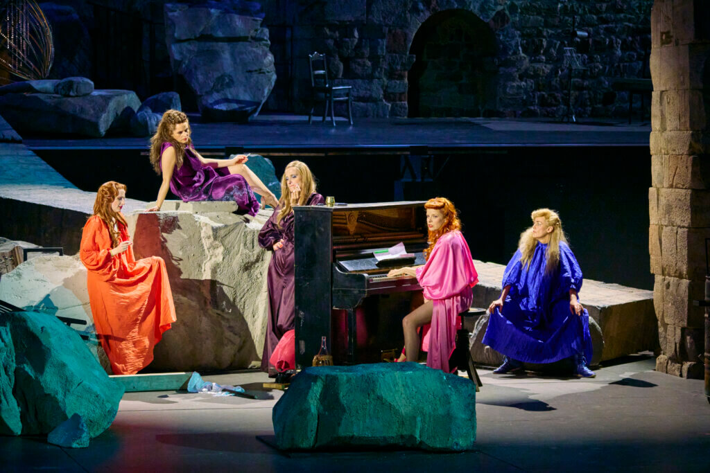 Aufführungsfoto von „Lysistrata“ von Amanda Lasker-Berlin nach Aristophanes bei den 75. Bad Hersfelder Festspielen. Eine Frau sitzt am Klavier, vier andere Frauen sitzen auf Felsblöcken drumherum.