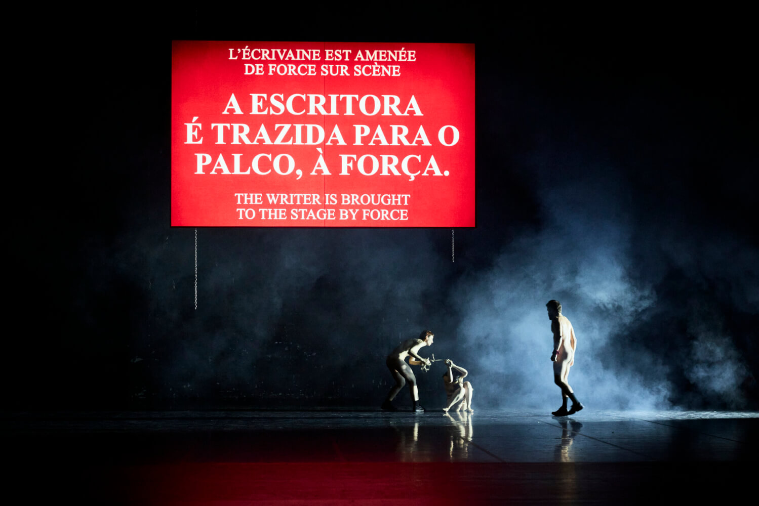 Auf der Bühne prangt ein großes Schild mit der Schrift: A Escritora é trazida para o palco, a Força. Darunter stehen drei Personen im Nebel.