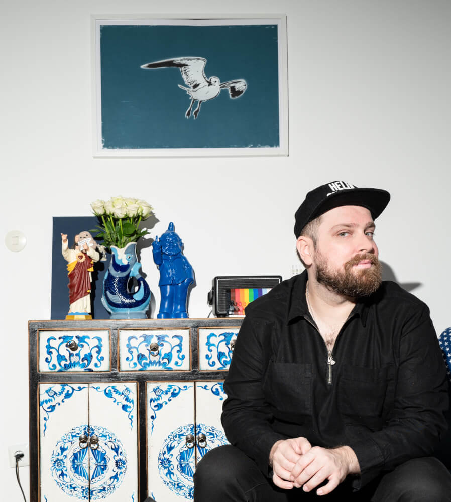 eine Person von vorne schräg im Profil vor einer Wand mit einem Bild und weiteren Gegenständen