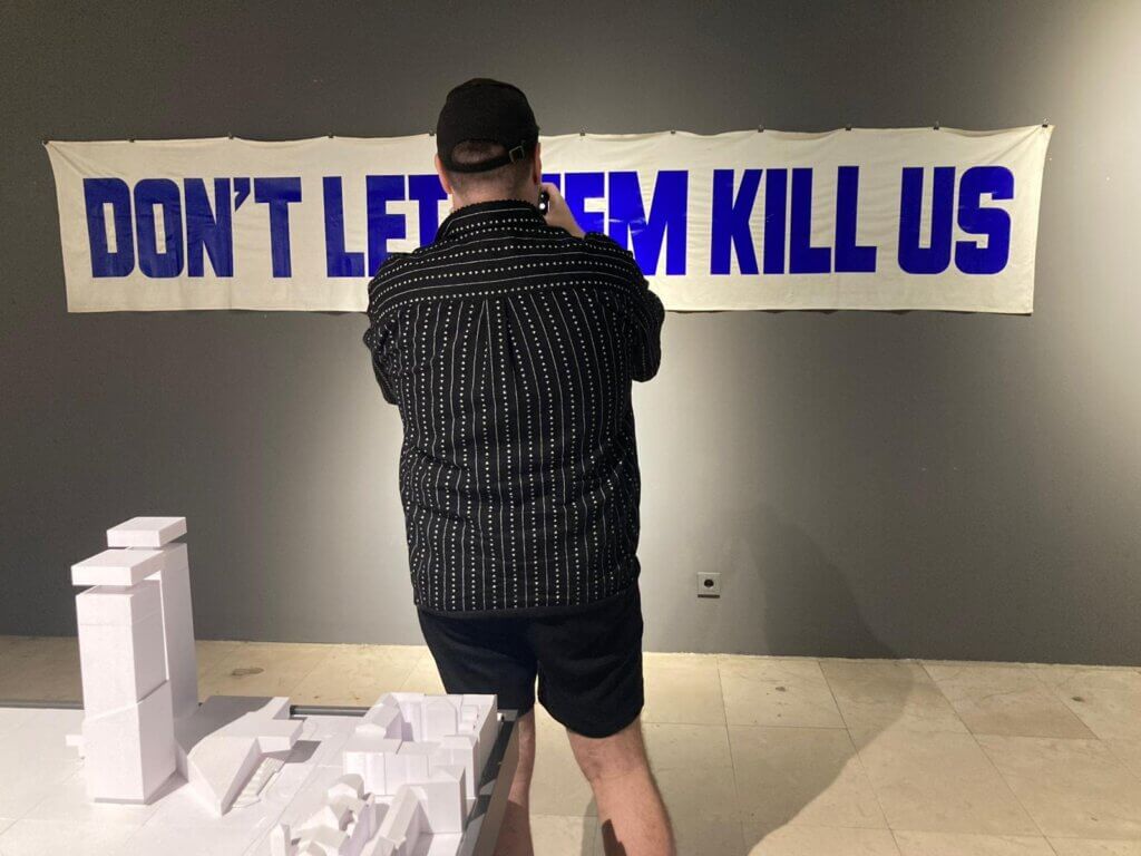 eine Person steht vor einem Banner auf dem „don't let them kill us“ steht in einem Museum