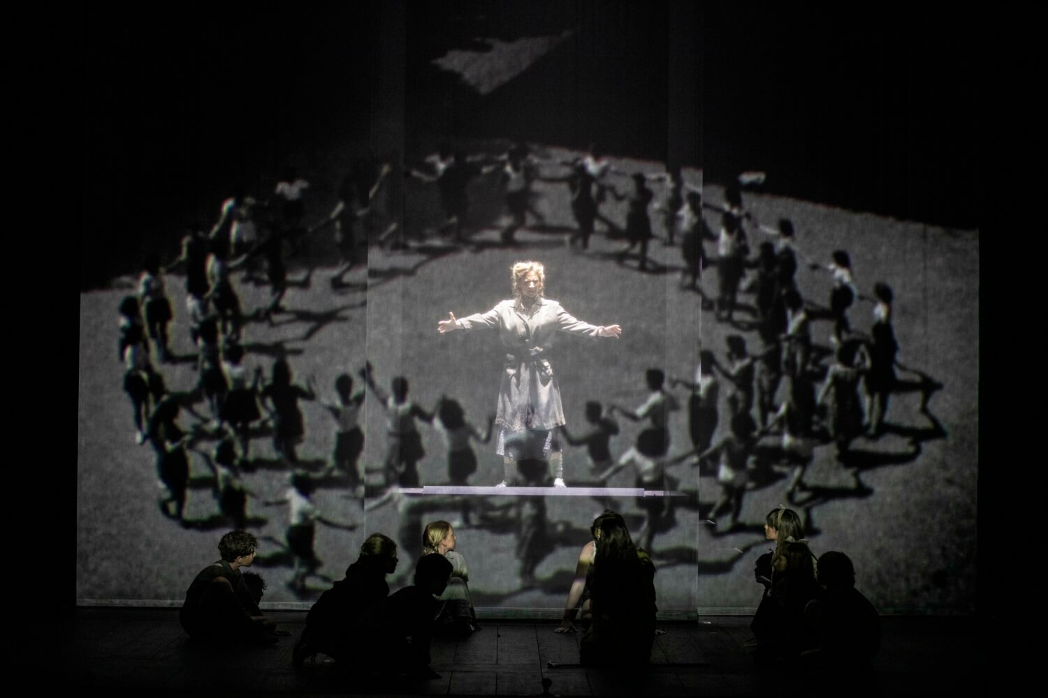 Aufführungsfoto von „Die Zauberflöte“ von Wolfgang Amadeus Mozart zur Eröffnung des Festival d’Aix-en-Provence. Auf einer Leinwand ist ein innerer und ein äußerer Kreis aus Menschen zu sehen. In der Mitte eine Frau. Im Bühnenvordergrund mehrere Menschen auf dem Boden sich zur Leinwand wendend.