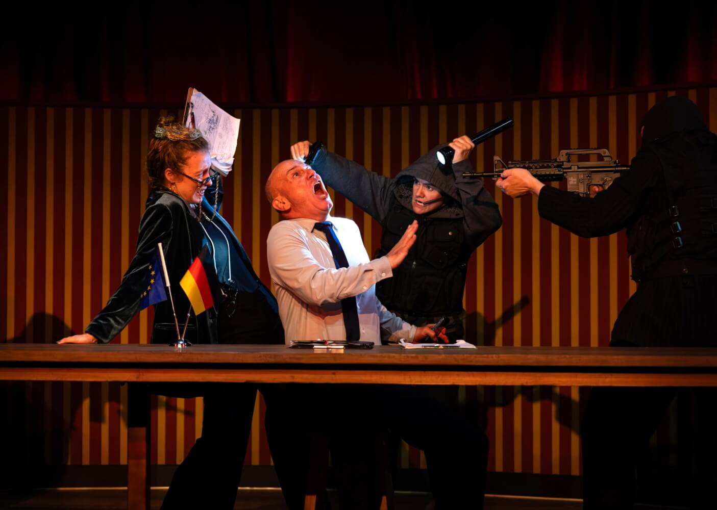 Aufführungsfoto von „Putsch“ von Alistair Beaton und Dietmar Jacobs am Metropoltheater München. Ein Mann im weißen Hemd und Krawatte sitzt an einem langen Holztisch. Zwei Personen in schwarzen Hoodies bedrohen ihn mit einer Pistole und einem Maschinengewehr und halten ihm eine Taschenlampe ins Gesicht. Hinter dem Mann steht eine Frau.