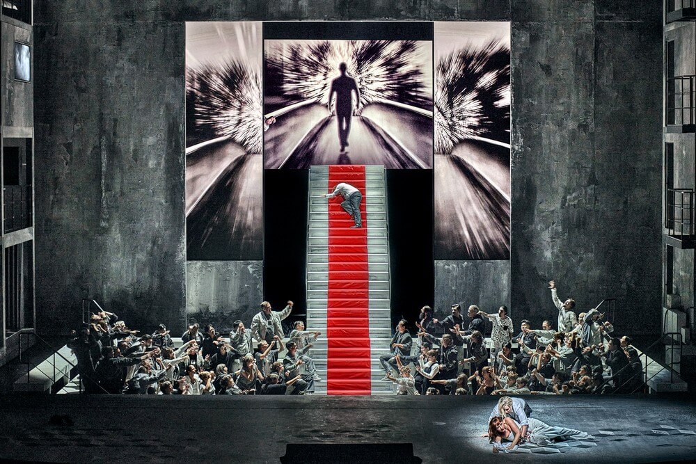 Aufführungsfoto von „Rienzi“ von Richard Wagner bei den 150. Bayreuther Festspielen. Menschenmassen links und rechts einer großen steilen Treppe, die zu einer großen Leinwand empor führt.