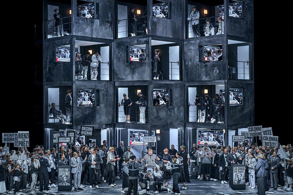 Aufführungsfoto von „Rienzi“ von Richard Wagner bei den 150. Bayreuther Festspielen. Eine Massenszene mit Plakaten und einem Wahlkampfstand. Alles in dunklen Grautönen gehalten. Dahinter ein Häuserblock mit grauen Wänden und Öffnungen.