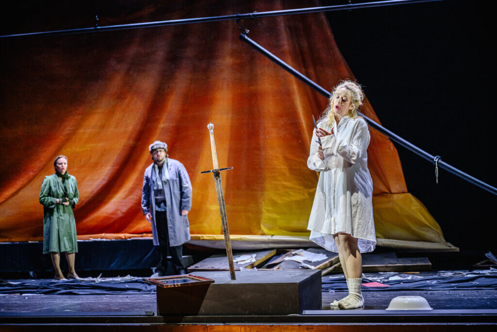 Lady Macbeth steht vorne rechts und ist wie im Wahn. Sie starrt ein Schwert in einem Stein an. Im Hintergrund stehen ihre Kammerzofe und ein Arzt und beobachten sie.