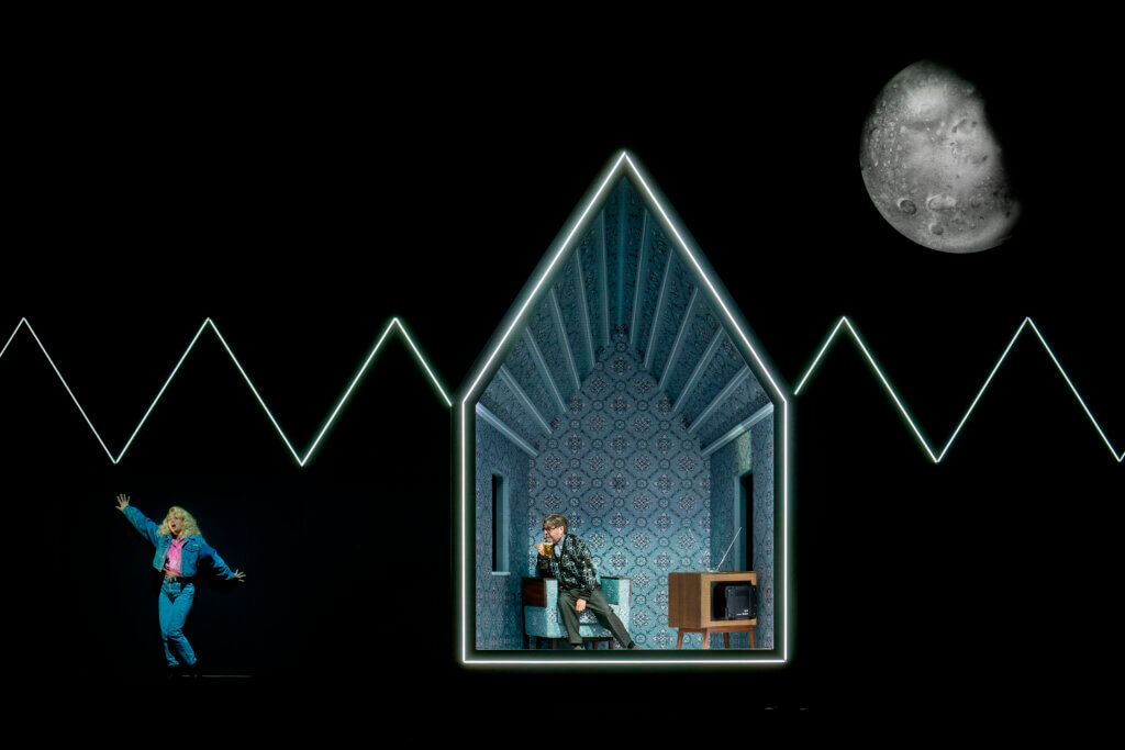 Die Bühne ist schwarz. Wie bei einer Zeichung ist im Hintergrund eine Linie eingezeichnet, die Bäume symbolisieren soll. Davor steht ein einfaches Haus, blau gegen den schwarzen Hintergrund ausgeleuchtet. Darin sitzt die Hauptfigur.