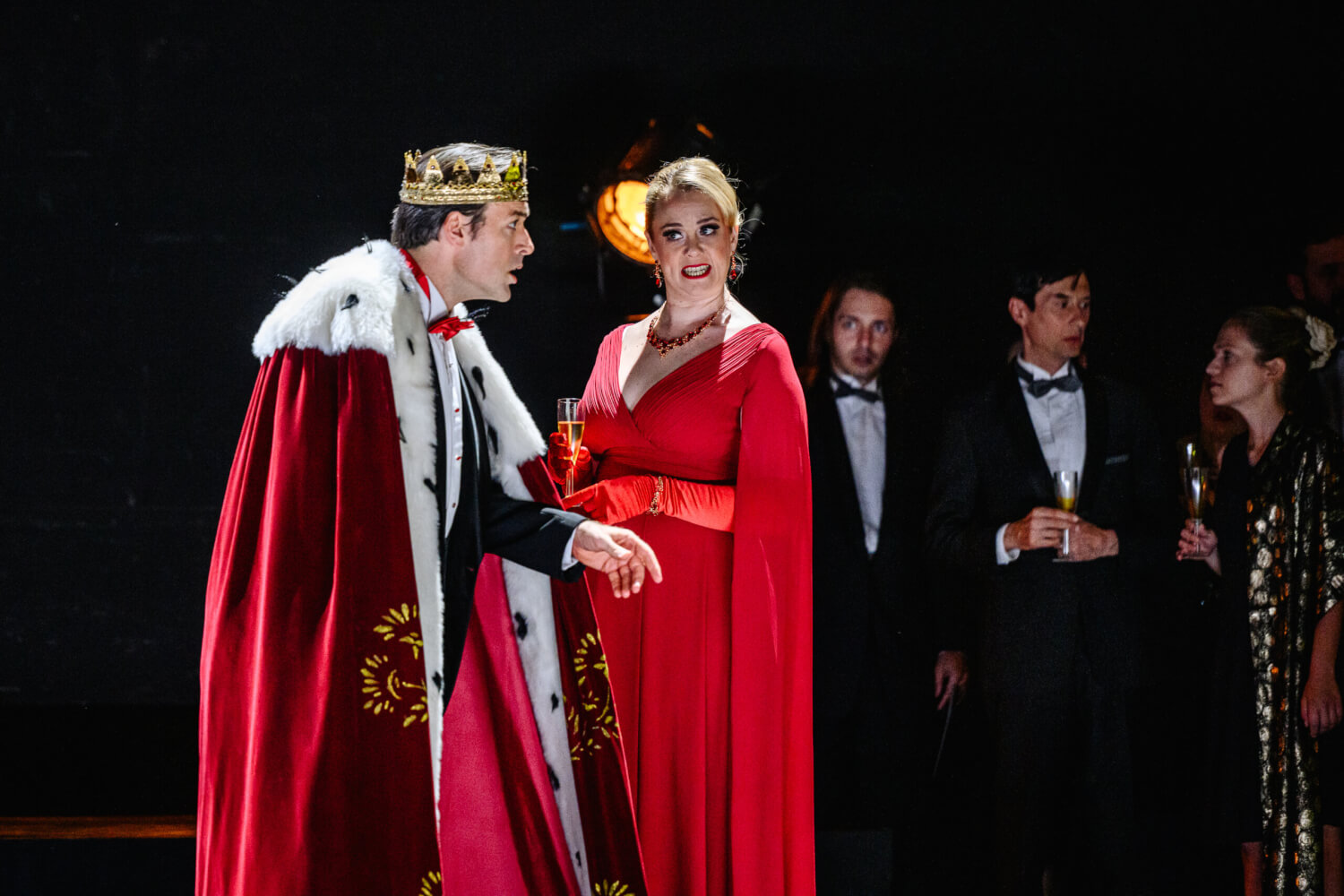 Thomas Weinhappel steht als Macbeth im roten Königsmantel und schaut seine Frau Lady Macbeth (Leah Gordon) an. Im Hintergrund steht ihre Gefolgschaft.