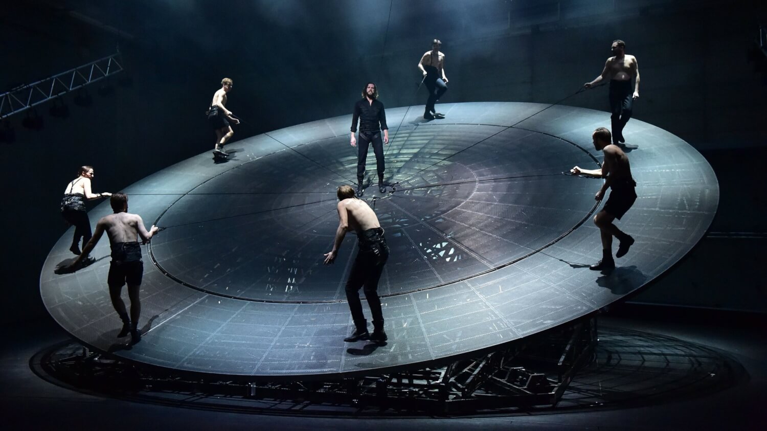 Aufführungsfoto von „Woyzeck“ am Theater Basel. Auf einer großen, schrägen Drehscheibe laufen sieben Männer oberkörperfrei am Rand der Scheibe, wie Uhrzeiger. In der Mitte der Scheibe steht ein Mann.