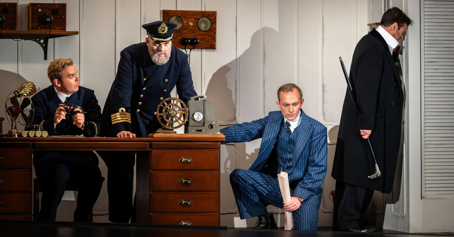 Aufführungsfoto von „Titanic“ am Theater Chemnitz. Ein Kapitän steht nach vorne gebeugt über einem Schreibtisch, an dem ein anderer Mann sitzt. Daneben kniet ein Mann im blauen Anzug. Rechts davon lehnt ein Mann stehend mit dem Kopf an der weißen Wand.