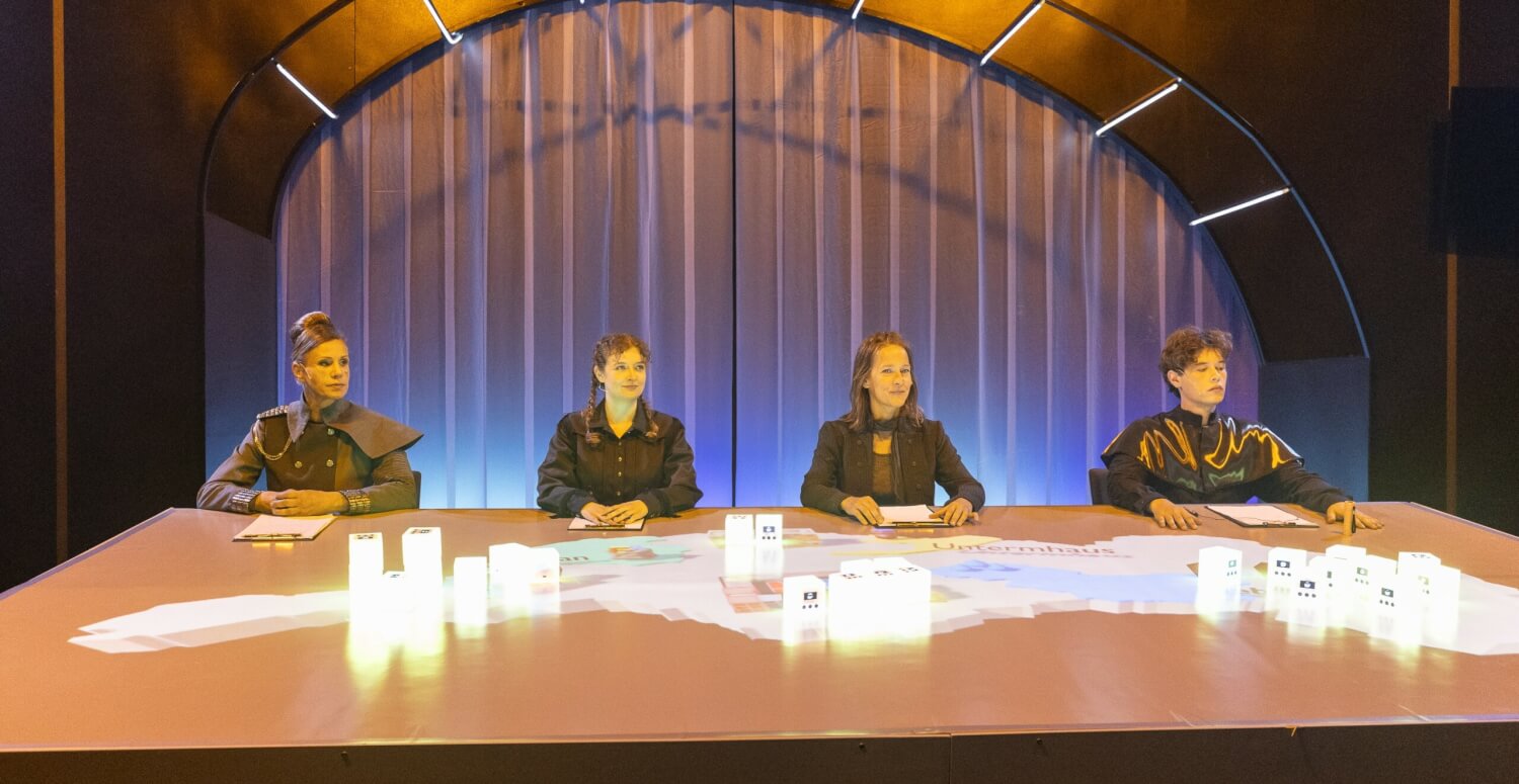 Aufführungsfoto von „√Mytopia“ von Sophie Oldenstein am Theater Altenburg Gera. Drei Frauen und ein Mann sitzen an einem großen Tisch, auf dem die Umrisse von Gera zu sehen sind, auf dem Tisch stehen weiß leuchtende Kuben.